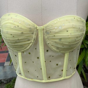 May include: A yellow mesh corset with gold star accents. The corset has a boned structure and a bustier style.