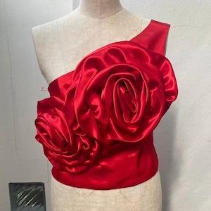 Puede incluir: Un top de satén rojo con un gran diseño de rosa tridimensional en el hombro.