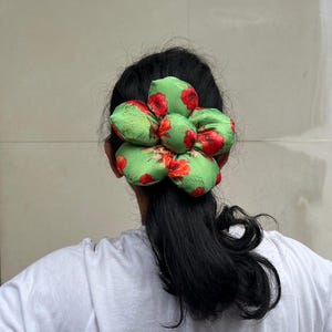 A green scrunchie with a floral pattern of red flowers and green leaves. The scrunchie is worn in the hair, and the fabric is gathered in a flower-like shape. The hair accessory is set against a neutral background.