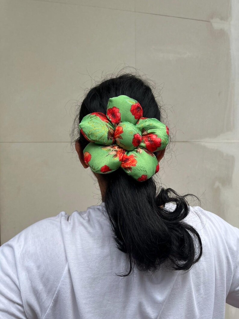 A green scrunchie with a floral pattern of red flowers and green leaves. The scrunchie is worn in the hair, and the fabric is gathered in a flower-like shape. The hair accessory is set against a neutral background.