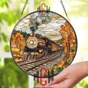 May include: Circular stained glass window art showing a vintage train scene. The artwork depicts a black and gold steam engine on a track, with autumn trees and a cloudy sky in the background. The window is suspended by a black chain.