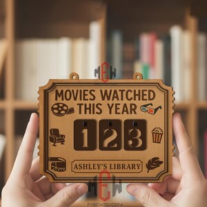 Op de afbeelding: Houten bord met de tekst "MOVIES WATCHED THIS YEAR" en de cijfers "123". Het bord heeft een decoratieve rand en bevat pictogrammen van een filmrol, 3D-bril, een stoel, popcorn, een filmrol en een blad. De tekst "ASHLEY'S LIBRARY" is ook opgenomen.