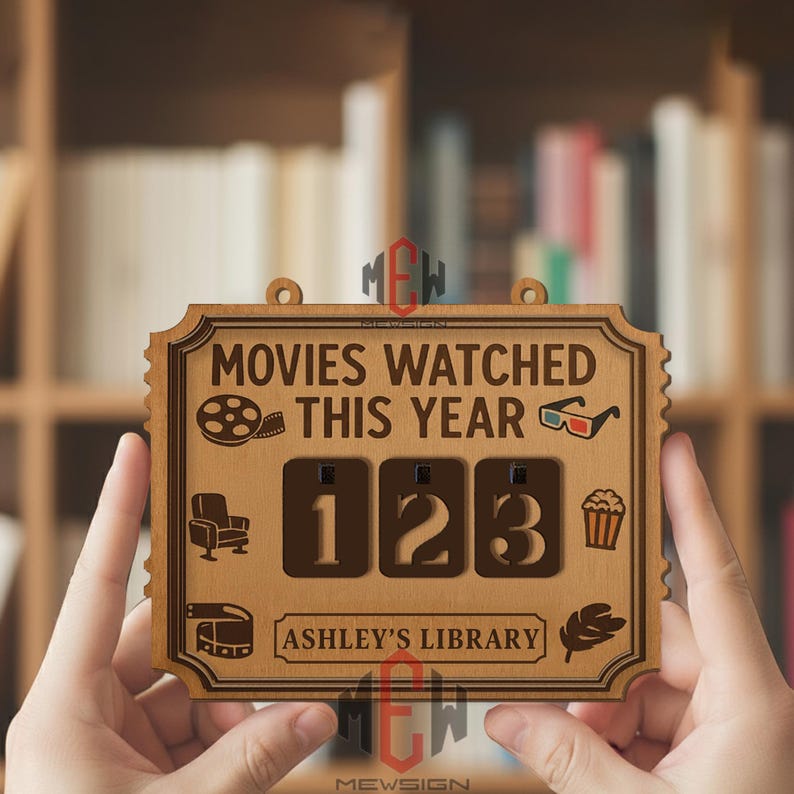Op de afbeelding: Houten bord met de tekst "MOVIES WATCHED THIS YEAR" en de cijfers "123". Het bord heeft een decoratieve rand en bevat pictogrammen van een filmrol, 3D-bril, een stoel, popcorn, een filmrol en een blad. De tekst "ASHLEY'S LIBRARY" is ook opgenomen.