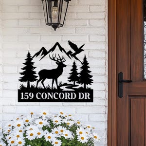 Peut inclure: Panneau d'adresse en métal noir représentant un cerf, des montagnes, des arbres et des oiseaux. Le panneau indique "159 CONCORD DR" en lettres blanches. Il est fixé sur un mur de briques blanches, près d'une porte en bois et d'une lanterne.