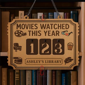 Op de afbeelding: Houten bord met de tekst "MOVIES WATCHED THIS YEAR" en het nummer "123". Het bord heeft filmgerelateerde iconen en de tekst "ASHLEY'S LIBRARY". Het hangt aan een ketting en wordt omringd door boeken.