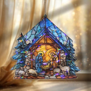 May include: A stained-glass Christmas ornament depicting the Nativity scene. The ornament features a blue roof, a warm-toned interior, and a detailed depiction of Mary, Joseph, and baby Jesus, surrounded by sheep. The ornament is designed to hang.