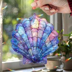 Peut inclure: Un capteur de soleil en forme de coquillage en vitrail aux tons de bleu, violet et rose. Le verre irisé capte la lumière, créant un effet scintillant. La pièce décorative est tenue par une main, mettant en valeur son design complexe et ses couleurs vives.