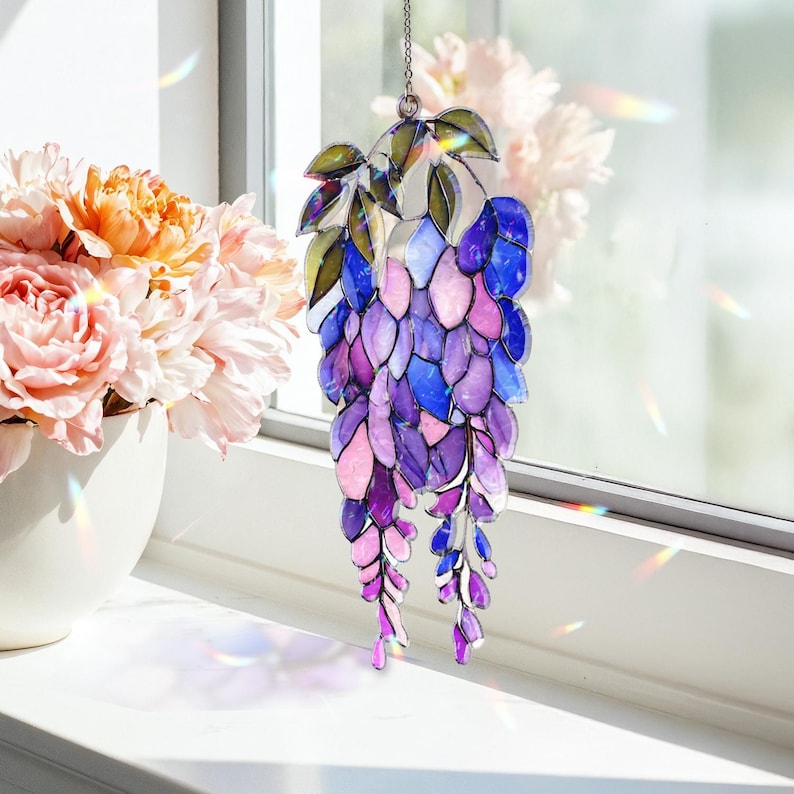 Pu&ograve; includere: Un acchiappasole a forma di glicine in vetro colorato con una catena per appenderlo. L'acchiappasole presenta una cascata di pezzi di vetro a forma di petalo viola, blu e rosa, con foglie verdi nella parte superiore. L'acchiappasole &egrave; appeso davanti a una finestra.