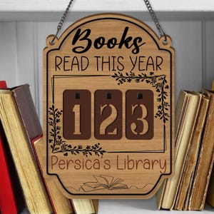 Könnte beinhalten: Holzschild mit dem Text "Books READ THIS YEAR" und einem Zähler, der "123" anzeigt. Das Schild hat ein dekoratives Blumendesign und den Text "Persica's Library". Eine Metallkette ist zum Aufhängen befestigt.