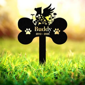 May include: Black metal pet memorial stake in the shape of a dog bone. The stake features a silhouette of a winged dog, flowers, and the name "Buddy" with the years 2012-2024. Two paw prints are on the bone.