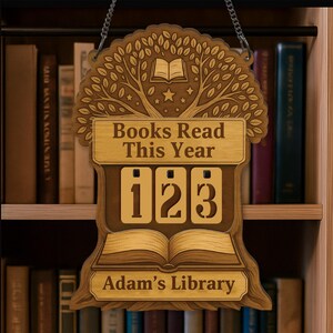 Könnte beinhalten: Holzschild mit dem Text "Gelesene Bücher dieses Jahr" und den Zahlen "123". Das Schild hat oben ein Baumdesign und unten die Worte "Adams Bibliothek". Es hängt an einer Kette vor einem Bücherregal.