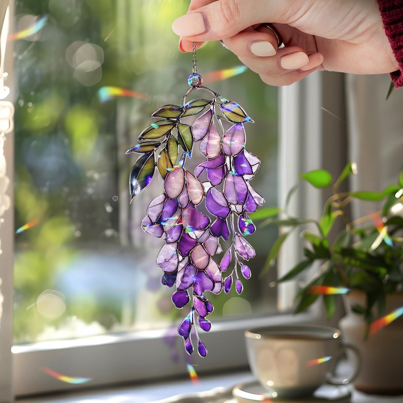 Pu&ograve; includere: Un acchiappasole a glicine in vetro colorato nei toni del viola, lavanda e verde. Il design floreale presenta foglie delicate e grappoli di fiori a cascata, sospesi da una catena d'argento. L'acchiappasole &egrave; tenuto davanti a una finestra.