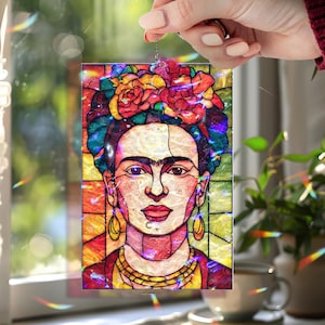 Puede incluir: Un atrapasoles de vidriera con un retrato de Frida Kahlo. La obra de arte presenta un tocado floral colorido, joyas de oro y una vibrante paleta de rojos, amarillos y verdes. El atrapasoles es sostenido por una mano.