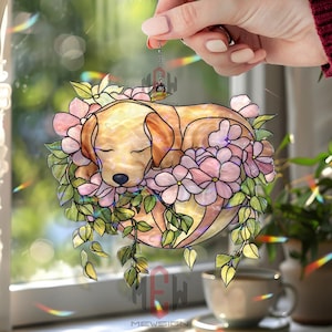 Puede incluir: Adorno de vidriera con un cachorro de Labrador dorado durmiendo, rodeado de flores rosas y hojas verdes. El cachorro es de color marrón claro con los ojos cerrados. El adorno cuelga de una cadena frente a una ventana.