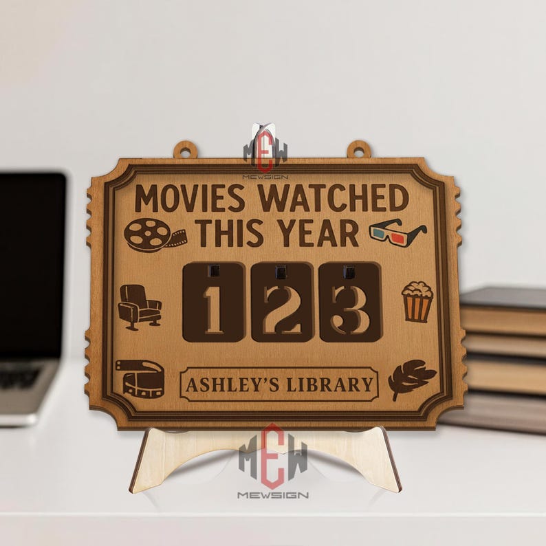 Op de afbeelding: Houten filmteller met de tekst "MOVIES WATCHED THIS YEAR" en de cijfers "123". Inclusief filmthema-iconen en de tekst "ASHLEY'S LIBRARY". De teller wordt ondersteund door een houten standaard.