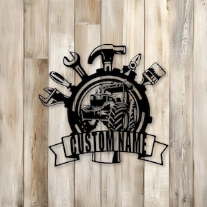 Könnte beinhalten: Schwarze Metall-Wandkunst mit einem Traktor in einem Zahnrad, umgeben von Werkzeugen wie Hammer, Schraubenschlüssel und Schraubendreher. Ein Banner darunter lautet "CUSTOM NAME". Auf hellem Holzgrund.