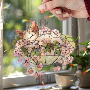 Könnte beinhalten: Ein Glasfenster-Katzenfänger, der eine schlafende Katze inmitten rosa Blüten und grünen Blättern zeigt. Die Katze ist hellbraun und cremefarben. Der Sonnenfänger hängt an einer Kette. Der Text "MEWSIGN" ist unten sichtbar.
