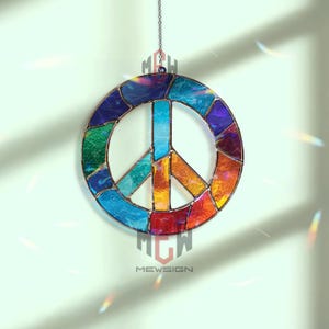 Puede incluir: Un atrapasoles de vidrio manchado con el símbolo de la paz, con una gama de colores del arcoíris, incluyendo azul, verde, naranja y rojo. El símbolo de la paz está suspendido por una cadena. La marca "MEWSIGN" está impresa en la parte inferior.