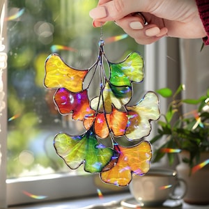 May include: A vibrant stained glass ginkgo leaf suncatcher. The leaves are a mix of yellow, orange, green, and purple, with a metallic frame and chain for hanging. The suncatcher is held up in front of a window.