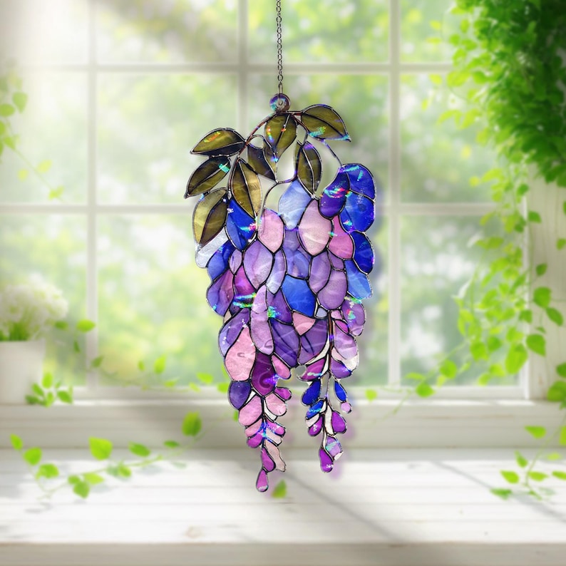 Pu&ograve; includere: Una decorazione a sospensione in vetro colorato a forma di glicine. Il pezzo presenta una cascata di pezzi di vetro a forma di petalo viola, blu e rosa, con foglie verdi e dorate. Appeso a una delicata catena.