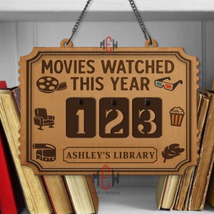 Op de afbeelding: Houten bord met de tekst "MOVIES WATCHED THIS YEAR" en de cijfers "123". Het bord heeft filmthema-iconen en de tekst "ASHLEY'S LIBRARY". Het hangt aan een ketting en is omgeven door boeken.