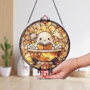 May include: A round stained glass window featuring a cartoon ghost in a bathtub reading a book. The window has a mosaic-like design with warm yellow and brown tones. The image is suspended by a chain.