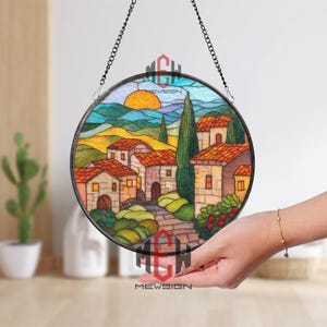 May include: A round stained glass panel depicting a village scene with a sunset. The artwork features houses with orange roofs, green trees, and a winding path. The panel is framed in black and suspended by a chain.