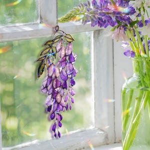 Pu&ograve; includere: Una decorazione a sospensione a vetrata a forma di glicine. Il pezzo presenta grappoli di fiori viola e lavanda con foglie verdi, sospesi a una catena. Lo stipite della finestra e un vaso di fiori sono sullo sfondo.
