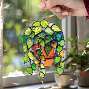 May include: Stained glass hanging plant decoration. The piece features a vibrant orange pot with green and yellow leaves. The leaves have a textured appearance and are arranged in a cascading pattern. The piece is held by a chain.