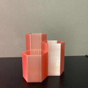 Può includere: Tre fioriere esagonali color corallo di diverse dimensioni sono disposte insieme. Le fioriere hanno una superficie strutturata e sono realizzate in materiale stampato in 3D. La disposizione suggerisce uno scopo decorativo o organizzativo.