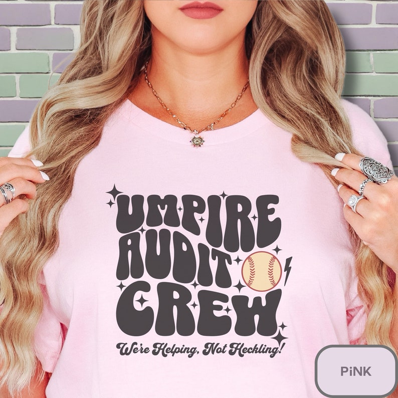 May include: Pink t-shirt with a retro-style graphic that says "Umpire Audit Crew" with a baseball and lightning bolt. The text below the graphic says "We're Helping, Not Heckling!"