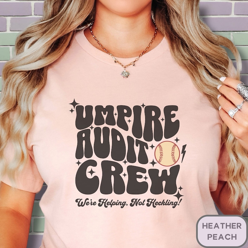May include: A pink t-shirt with the text "Umpire Audit Crew" in a retro font. A baseball graphic is included with the text "We're Helping, Not Heckling!"