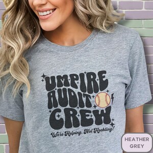 May include: Heather grey t-shirt with a black graphic that says "Umpire Audit Crew" with a baseball and lightning bolt. The text is in a retro font style. The shirt also has the text "We're Helping, Not Heckling!" in a smaller font.