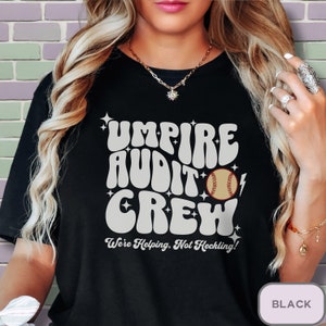May include: Black t-shirt with a white graphic that says "Umpire Audit Crew" in a retro font. A baseball and lightning bolt are included in the design. The text "We're Helping, Not Heckling!" is below the graphic.