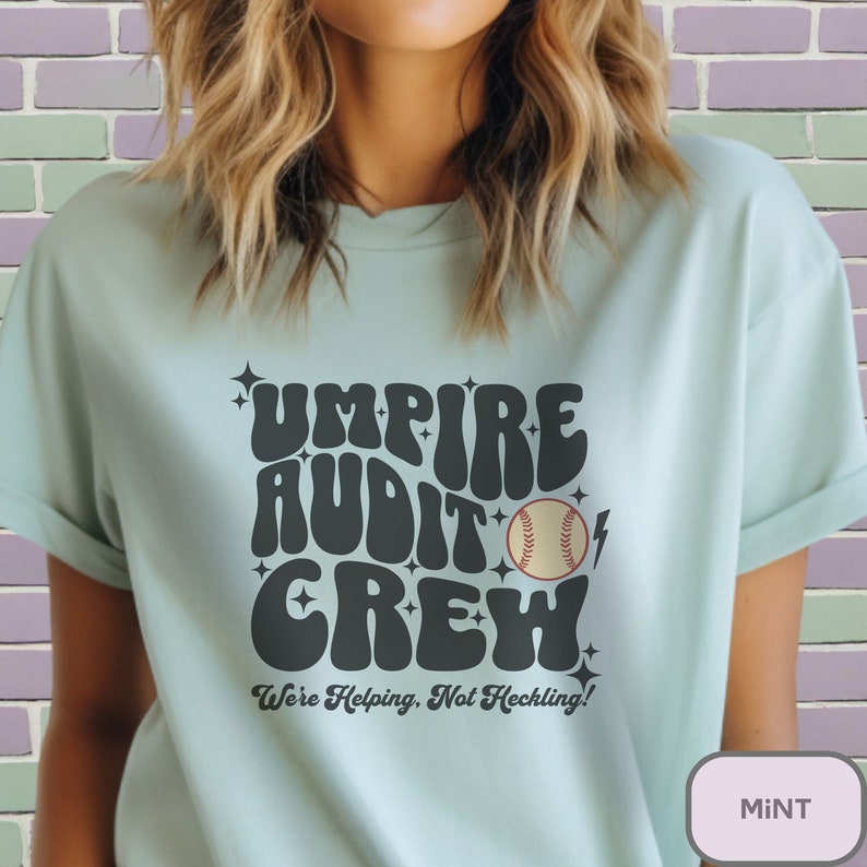 May include: A light blue t-shirt with a black graphic that says "Umpire Audit Crew" in a retro font. A baseball is included in the graphic. The text below the graphic says "We're Helping, Not Heckling!"
