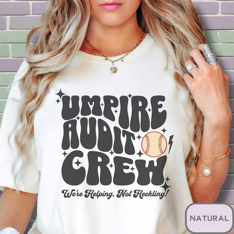 May include: White t-shirt with a retro-style graphic that says "Umpire Audit Crew" with a baseball graphic and the text "We're Helping, Not Heckling!"