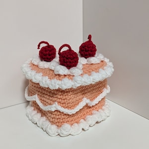 May include: A crocheted cake-shaped tissue box cover with white frosting and three red cherries on top.