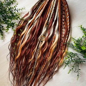 Puede incluir: Un conjunto de rastas en tonos marrones, naranjas y rubios. Las rastas tienen puntas onduladas y algunas están trenzadas. Algunos mechones están adornados con cuentas y un dije circular. Ramitas de hojas verdes se colocan alrededor de las rastas.