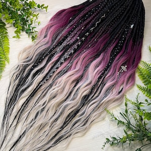 Könnte beinhalten: Ein Set aus schwarzen, lila und hellrosa Kunsthaar-Dreadlocks. Die Dreadlocks sind mit silbernen Akzenten und Charms geflochten. Die Enden der Dreadlocks sind hellblond. Die Dreadlocks sind auf einer hellen Holzoberfläche mit grünem Laub angeordnet.