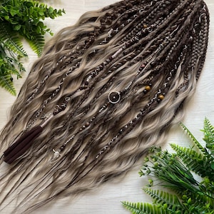 May include: A collection of brown and blonde synthetic dreadlocks, adorned with silver charms, wooden beads, and white stitching. The dreadlocks have a wavy texture and a dark brown tassel. The hair is displayed on a light wood surface, with green foliage.