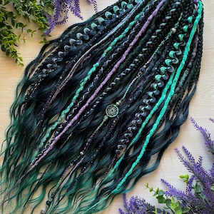 Può includere: Una collezione di dreadlock neri e verde acqua scuro con estremità ondulate. I dread sono adornati con accenti turchesi, viola e argento, tra cui perline e un ciondolo acchiappasogni. Lo stile è disposto su una superficie chiara con rametti di lavanda.