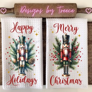 May include: Two white waffle-weave kitchen towels featuring nutcracker graphics. The left towel says Happy Holidays with a nutcracker, pine branches, and pinecones. The right towel says Merry Christmas with a nutcracker, pine branches, and red berries.