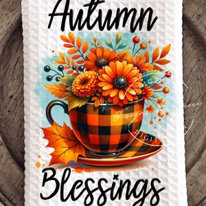 May include: A white waffle-weave kitchen towel featuring a vibrant autumn-themed design. The artwork includes a teacup with orange and black plaid, filled with orange flowers and fall foliage. The words "Autumn Blessings" are written in black script.