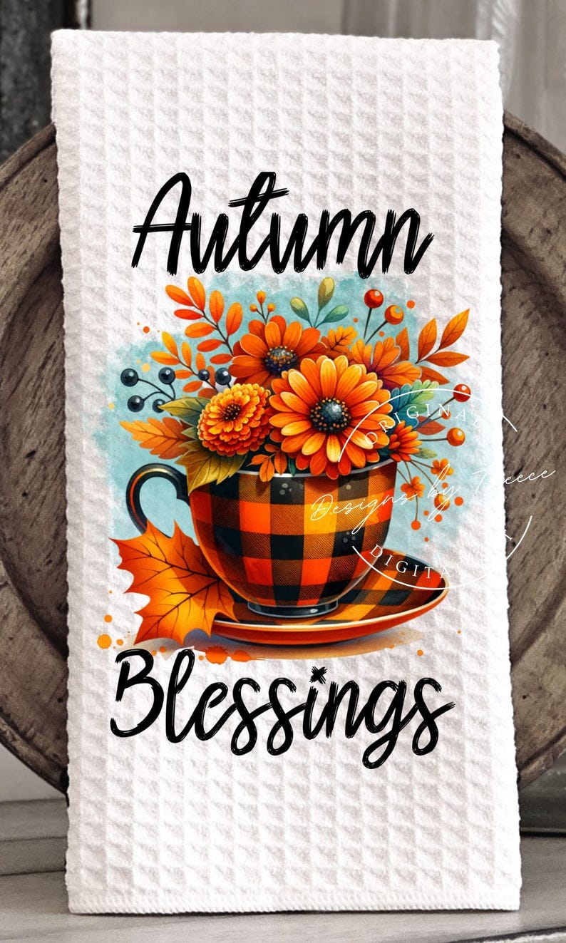 May include: A white waffle-weave kitchen towel featuring a vibrant autumn-themed design. The artwork includes a teacup with orange and black plaid, filled with orange flowers and fall foliage. The words "Autumn Blessings" are written in black script.