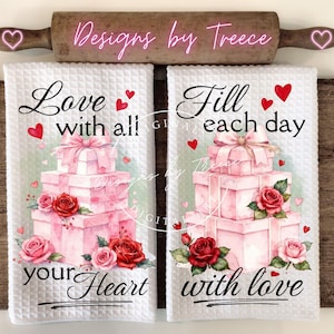 Op de afbeelding: Twee witte keukenhanddoeken met wafelstructuur met de zinnen "Love with all your Heart" en "Fill each day with love". Elke handdoek heeft een stapel roze cadeau dozen, rode rozen en harten. Bovenop ligt een houten deegroller.