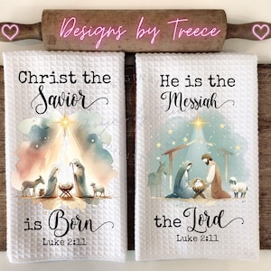 May include: Two white waffle-weave kitchen towels featuring watercolor nativity scenes. The left towel reads Christ the Savior is Born Luke 2:11. The right towel reads He is the Messiah the Lord Luke 2:11. Both display figures of the holy family.