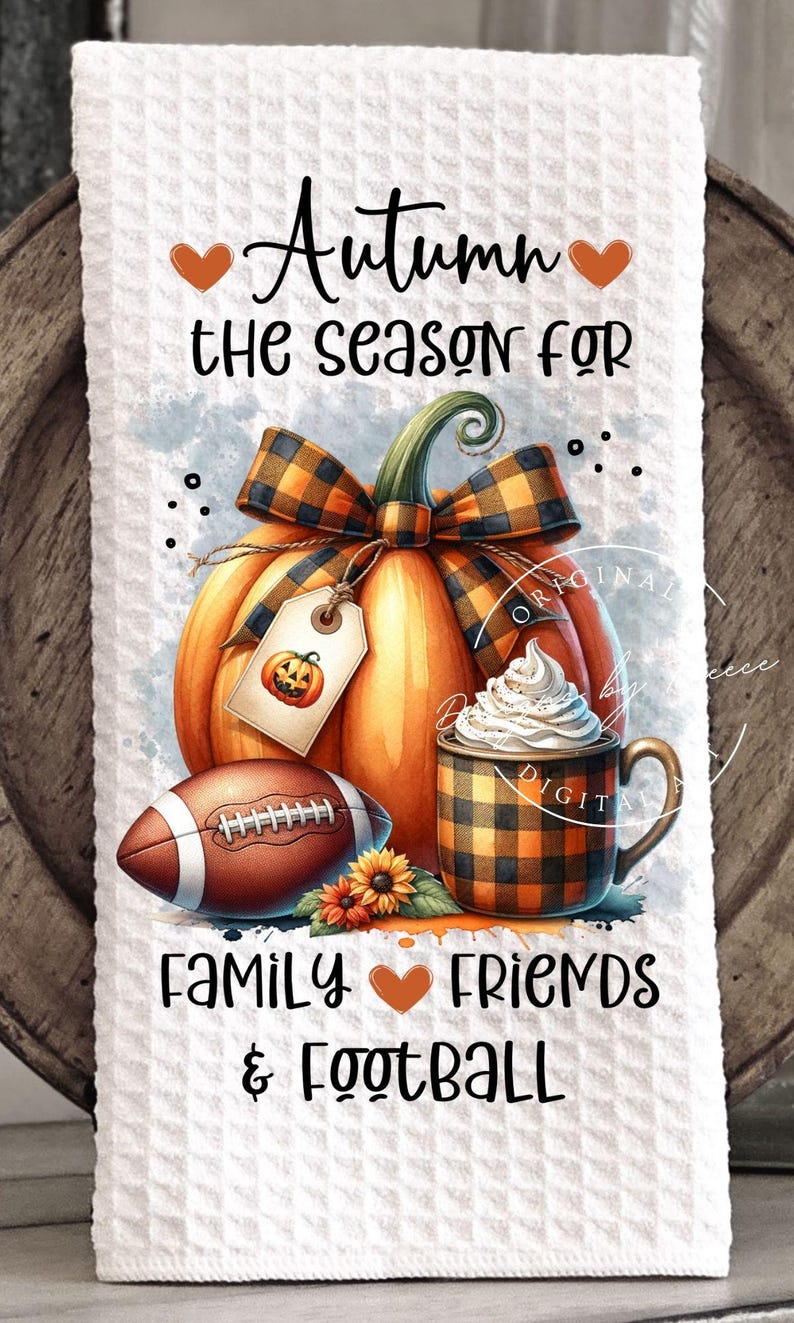 May include: White kitchen towel with an autumn-themed design. Features a large orange pumpkin with a plaid bow, a football, and a mug of whipped cream. Text reads "Autumn the season for family, friends & football."