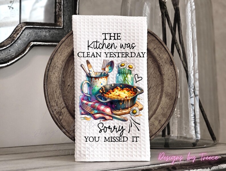 May include: White kitchen towel with a waffle weave texture. The towel features a colorful illustration of kitchen utensils, a casserole dish, and a jar of daisies. Text on the towel reads "THE kitchen was clean yesterday, Sorry YOU MISSED IT."