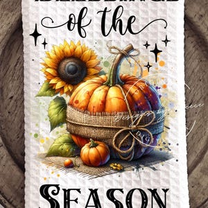 May include: A white waffle-weave kitchen towel with a fall-themed design. The image features a sunflower, pumpkins, and the text "BLESSINGS of the SEASON" in black script. The pumpkins are wrapped in burlap.