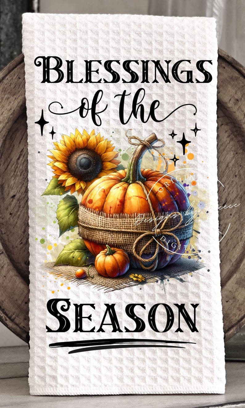 May include: A white waffle-weave kitchen towel with a fall-themed design. The image features a sunflower, pumpkins, and the text "BLESSINGS of the SEASON" in black script. The pumpkins are wrapped in burlap.